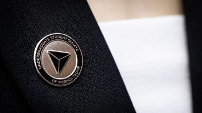 A metal pin affixed to a black jacket over a white shirt. The pin is metallic with a black background and has a three-sided pyramid design in the center. "Undergraduate Student Senate of Virginia Tech" is engraved around the outside border of the pin.