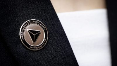 A metal pin affixed to a black jacket over a white shirt. The pin is metallic with a black background and has a three-sided pyramid design in the center. &quot;Undergraduate Student Senate of Virginia Tech&quot; is engraved around the outside border of the pin.