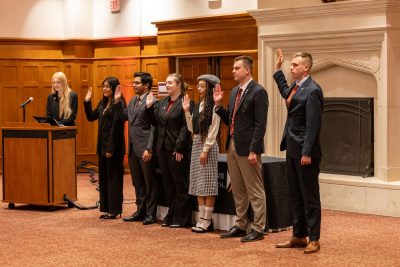 Several students in formal wear stand side by side with their right hands raised. Another student stands at a lectern with a microphone. The background is a wood paneled wall and a large stone fireplace.